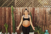 Pregnant woman in black top and leggings standing in garden with cactuses and wood fence, highlighting maternity theme.